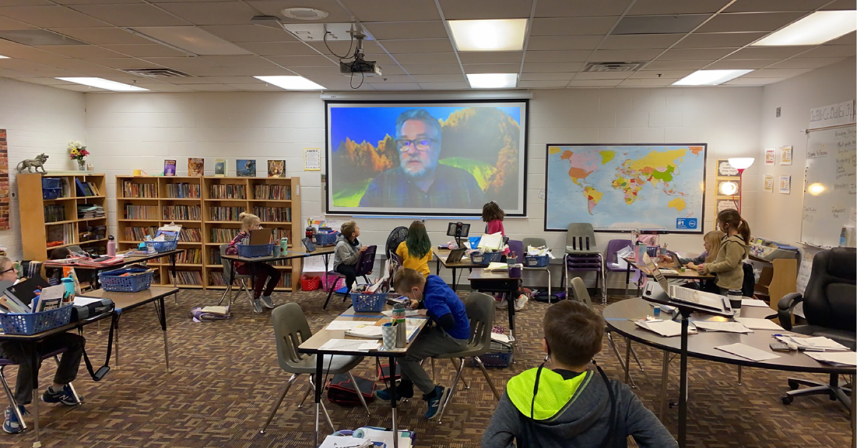 Students in a classroom participating in a virtual author visit with Mark Cheverton projected on a screen.